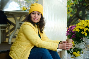 Sarı şapkalı ve ceketli çekici bir kadının yan portresi, kağıt bardakta aromalı kahve, tencere kenarında renkli çiçeklerin yanında oturuyor, güneşli sonbahar gününün tadını çıkarıyor.