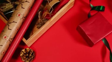 Kırmızı ambalaj kağıdına sarılı yeni yıl hediyesi, parlak yeşil bir kurdele ve şenlikli süsler, altın çam kozalakları, kırmızı yüzeyde ahşap bir sandıkta keten bir ip.