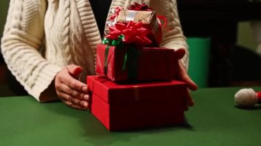 Ön tarafı kapatın. Bej renkli, sıcak kazaklı bir kadın, yeşil bir masaya Xmas hediyelerini bire bir koyuyor. Mutlu Noeller ve mutlu yıllar konsepti. Boks Günü. Kutlama etkinlikleri için hediye paketleme