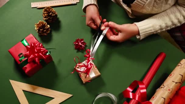 はさみを使用して女性の手のトップビューは クリスマスの贈り物を結びながら ストリップに白い装飾リボンを切断します 紙を包む 緑の表面に装飾として松のコーン 新年の準備 — ストック動画