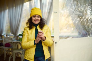 Çekici kıvırcık kadın, yeşil ceketli, parlak sarı şapkalı ve ceketli, ellerini sıcak aromalı kahve ile ısıtıyor kağıt bardakta, güzel dişlek bir gülümsemeyle kameraya gülümsüyor