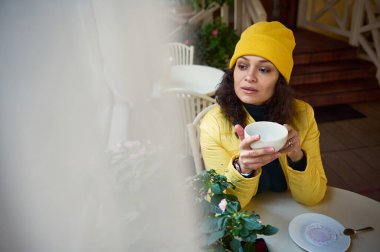 Ön plandaki bulanık perdelerin ardından sarı ceketli, şapkalı, çok ırklı, esmer bir kadına dikkatlice pencereden dışarı bakarken, rahat bir kahve dükkanında kahve molası veriyor.