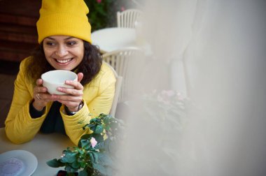 Ön plandaki bulanık perdelerin arasından sarı ceketli ve şapkalı hoş, çekici bir esmer kadının kahve molasının keyfini çıkarırken sıcak bir yudum sıcak içki içişini izleyin.