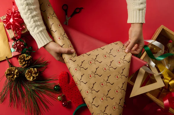 stock image Top view of crafts womans hands, rolling out a wrapping gift paper with deer pattern, over a red surface, with laid out wrapping materials, decorative ribbons and bows, pine cones as Christmas decor