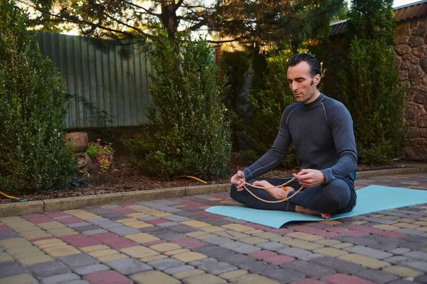 stock image Peaceful man yogi sitting in lotus pose on a fitness mat, meditating with rosary beads outdoors at sunset. Yoga practice. Meditation. Enlightenment. Body conscious. Active and healthy lifestyle