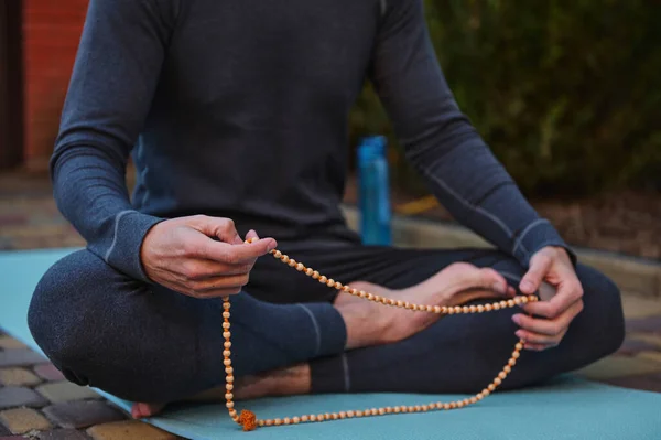 Cropped view of a yogi sitting on a fitness mat, meditating with rosary beads while practicing yoga outdoors. Mindfulness. Spiritual growth. Awakening. Enlightenment. Meditation. Body care