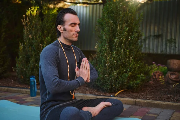 stock image Peaceful man yogi sitting in lotus pose on fitness mat, keeping hands palms together while meditating with rosary beads outdoors at sunset. Yoga practice. Meditation. Body conscious. Healthy lifestyle