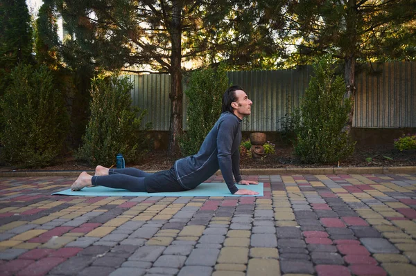 Stock image Side view a male athlete in gray activewear, a determined sportsman stretches his body on a yoga mat while practicing bodyweight training outdoors. Fitness. Stretching. Sport. Active lifestyle