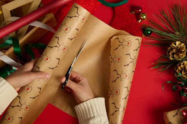 Top view of a woman in beige sweater, using scissors, cuts the wrapping gift paper with deer pattern over a red background, surrounded by wrapping materials and Christmas decorative items. Boxing Day