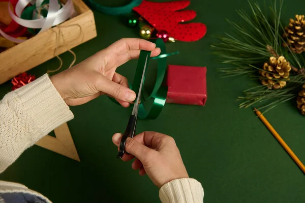 Top view of a woman in beige sweater, using scissors, cutting shiny green tape into strips for decorating the Christmas gift, wrapped in red decorative paper over a background with wrapping materials