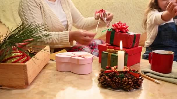 テーブルの上でプレゼントやラッピングの装飾的な材料に焦点を当て 照明付きのキャンドルは リビングルームでクリスマスプレゼントを梱包する家族の背景にあります 幸せな魔法のクリスマスの雰囲気 — ストック動画