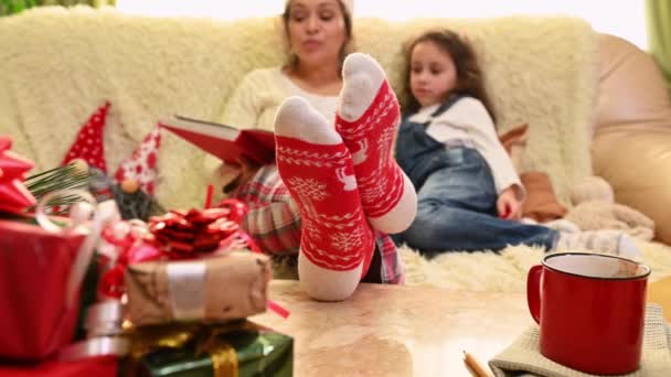 幸せな愛する母親のぼやけた背景に 彼女の愛らしい小さな娘におとぎ話を読んで 家で一緒に冬の休日を楽しんで クリスマスパターンの赤い靴下に焦点を当てます — ストック動画