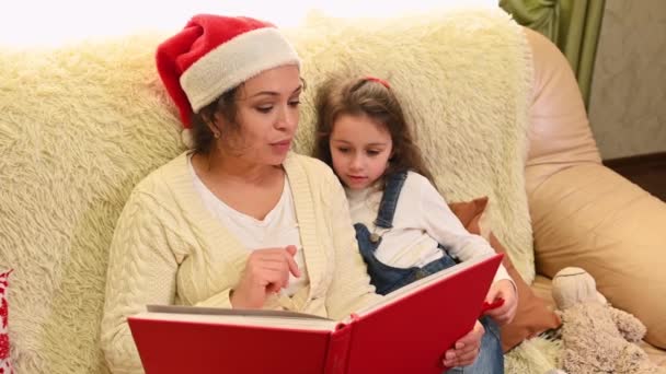 多民族的漂亮女人 穿着米黄色毛衣 头戴桑塔帽的可爱妈妈 坐在她可爱的小女儿旁边舒服的沙发上 给她读童话 家人的圣诞假期 — 图库视频影像