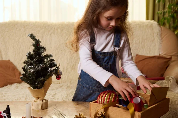 穿着白色衬衫和蓝色斜纹棉布工作服的漂亮小女孩 站在写日记的大理石桌旁 桌上摆放着木箱里的圣诞玩具 在舒适的家中享受新年和圣诞节的准备工作 — 图库照片