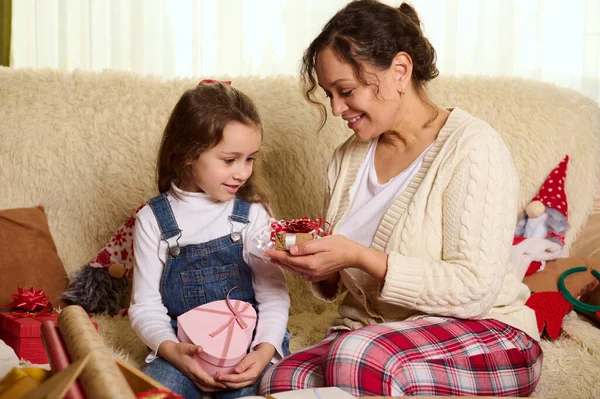 stock image Happy family relationships. Beautiful woman in beige sweater and red checkered trousers - a loving mom and her cute daughter exchanging Christmas presents, sitting together on sofa in the living room