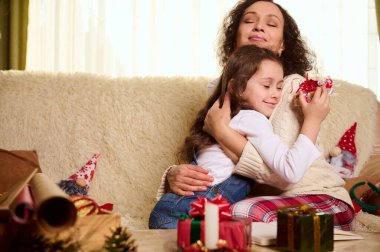 Sevimli küçük bir kız, sevgili kızı annesinin omuzlarına yaslanıp ona sarılıyor ve güzel bir Noel hediyesi alıyor. Öndeki masada güzel hediyeler var. Mutlu tasasız çocukluk