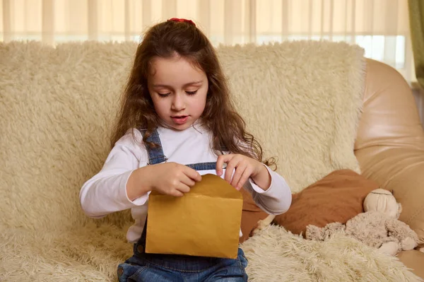 Charming Caucasian child, a 5 years little girl puts a letter to Santa Claus in a golden envelope, with her Christmas wishes. Magical festive winter atmosphere. New Years preparations. Happy childhood