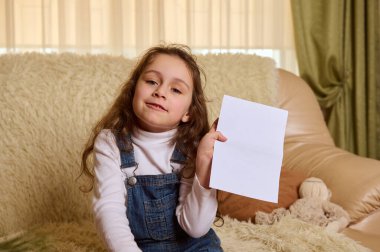 Küçük beyaz mutlu kız çocuğu, kameraya kurnazca gülümsüyor, içinde metin için fotokopi reklamı olan boş bir kağıt çarşaf gösteriyor, rahat bir evde rahat bir kanepede oturuyor.