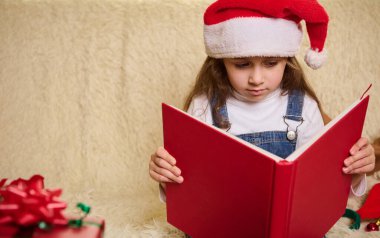 Noel Baba şapkalı şirin bir kız çocuğunun portresi, üstü örtülü kırmızı bir kitap, sıcacık bir evde Noel süsleriyle peri masalları okuyor. Reklam metni için reklam alanını kopyala. Yeni Yıl