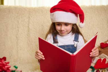 Çekici beyaz çocuk, Noel Baba şapkalı sevimli küçük kız, elinde kırmızı bir ciltli kitap, peri masalları okuyor, kanepede oturuyor, Noel süsleri ve hediyelerle çevrili, rahat bir evde.