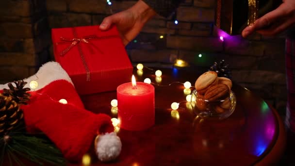 キャンドルが灯された木製のテーブルの上に幸せなプレゼントを積み重ねる女性の手の閉じる 自家製おいしい焼き菓子 サンタの帽子 装飾的な松のコーンと輝くガーランド メリークリスマス — ストック動画