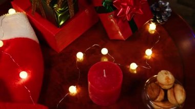 Noel hediyeleri, Noel Baba şapkası, fırınlanmış kurabiyeler ve parlak toplardan oluşan parlak çelenklerle dolu ahşap bir masada kırmızı dekoratif mum. Kış tatillerinde sihirli atmosfer