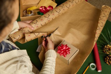 Overhead view of an unrecognizable woman, using scissors, cuts the decorative wrapping paper for packing gift for Christmas, New Year or other celebration event. Handsworkart. Boxing Day. Diy presents clipart