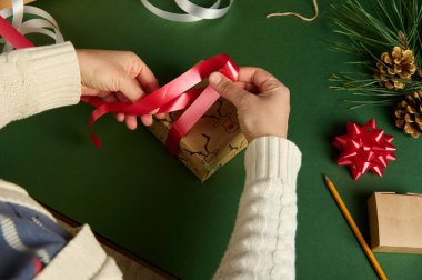 Üst görüşlü kadının elleri, kırmızı parlak bir kurdele kullanarak geyik desenli bir kağıda sarılı bir hediyeyi bağlıyor. Noel ve yeni yıl hazırlıkları. Diy. Yaratıcı bir fikir. El işi sanatı. Boks Günü. Yaratıcılık