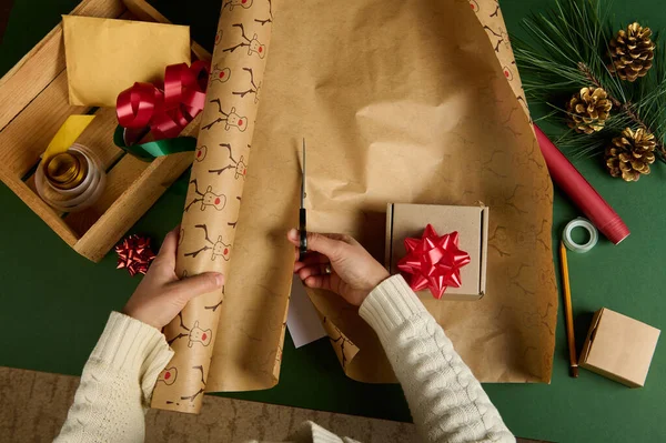 Top view woman in warm beige sweater, using scissors, cuts a wrapping paper with deer pattern, for packing gift for Christmas, New Year or other celebration event. Boxing Day. Diy present. Copy space