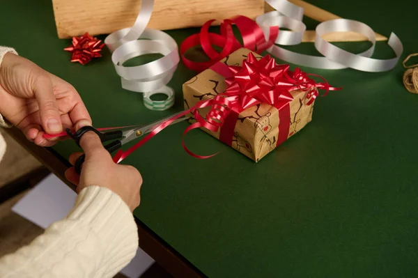 用剪刀对女人的手进行特写 用红色发亮的缎带剪成条状 用来在包装圣诞礼物时做漂亮的装饰品 并在绿色的背景上贴上广告 — 图库照片