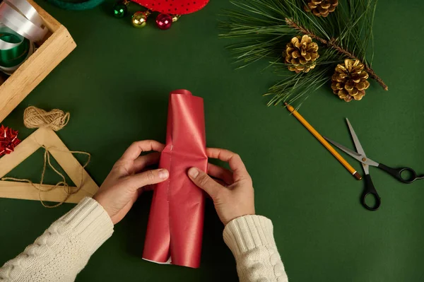 一个女人在绿色背景上用红色装饰纸包装一个小礼物 包装材料和金松果作为圣诞装饰的头像 迪伊的礼物节礼日 — 图库照片