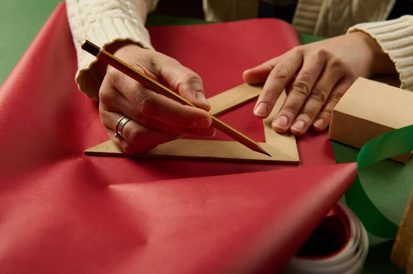 女人的手 用木制蜡笔和三角形尺子画在红色包装纸上 用来计算情人节 圣诞节或新年礼物的包装量 — 图库照片
