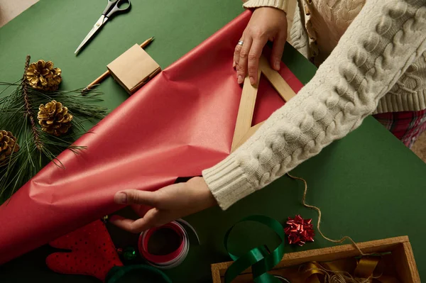 木製の三角形の定規を使用して認識できない女性の上のビューでは 緑の背景にクリスマスや新年の贈り物を梱包するために必要な赤い包装紙の量を計算します ボクシングの日 — ストック写真