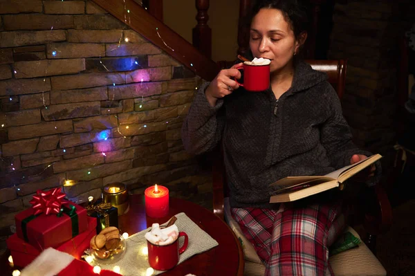 Relaxed multi ethnic beautiful woman, dressed in fluffy warm home clothes, drinking hot chocolate with marshmallows in a cozy home interior for Christmas. Winter holiday. December 25th. Happy New Year