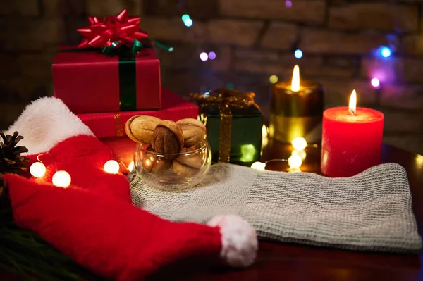 Home interior at Christmas with glowing lights on Santa hat, happy presents, homemade pastries and lit candles on festive table, on the brick wall background with hanging illuminated colorful garland