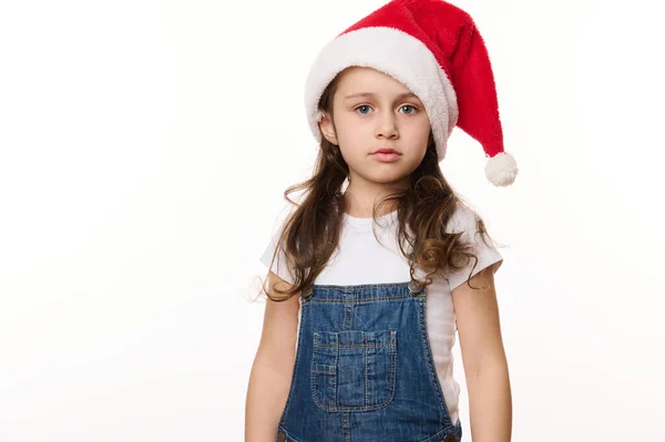 Isolated portrait on white background of a fancy cute Caucasian little girl in santa hat, in anticipation of Christmas holidays. Pure joy. Winter spirit. New Year party. Copy spacefor advertising text