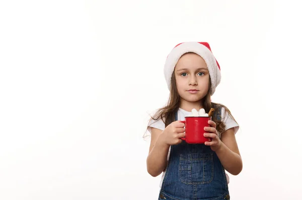 Isolated portrait on white background of a happy little girl in Santa hat, enjoying a delicious sweet hot chocolate drink with marshmallows. Christmas. Winter holidays concept. Copy ad space for text