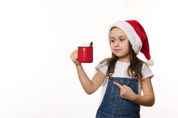 Isolated portrait on white background of a lovely baby girl in Santa, hat pointing at red cup of hot cocoa drink with marshmallows in her hands. Merry Christmas. Winter holiday atmosphere. Copy space