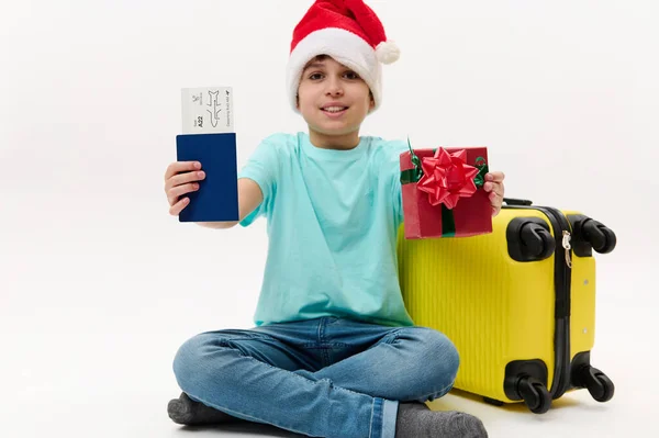Focus on a flight ticket in a passport document and Xmas gift in hands of a preteen boy in Santa hat, with bright yellow suitcase. Travelling at winter holidays. Tourism. Advertising for travel agency