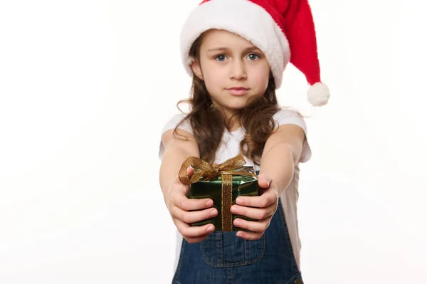 白人可爱的女婴 一个戴着圣诞老人帽和蓝色斜纹棉布工作服的小孩 手里拿着一件快乐的圣诞礼物 看着相机 被白色隔离 圣诞及新年准备工作 图库图片