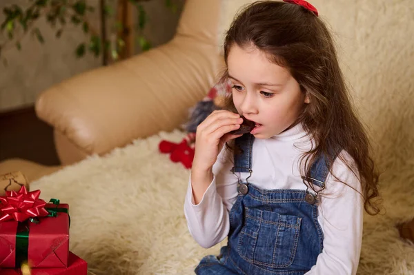 Beyaz tenli şirin çocuk, güzel uzun saçlı, güzel bir kız bebek beyaz gömlek ve mavi kot pantolon giyiyor, evde koltukta Noel hediyeleriyle otururken çikolata yiyor.