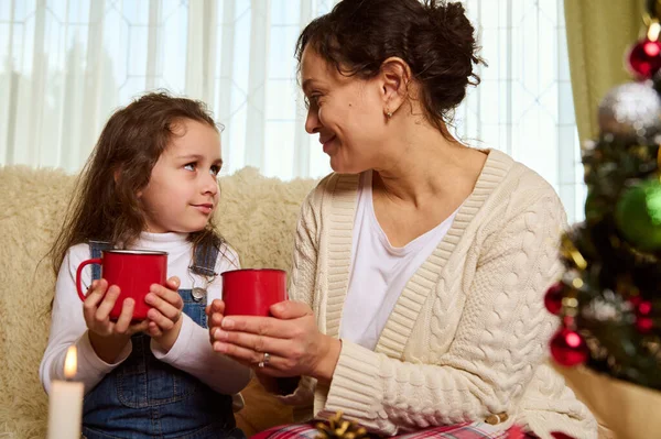 Beautiful multi-ethnic family of a loving mother and her lovely daughter, with mugs of hot chocolate drink, enjoying happy moments together at Christmas holidays in cozy home interior. December 25th