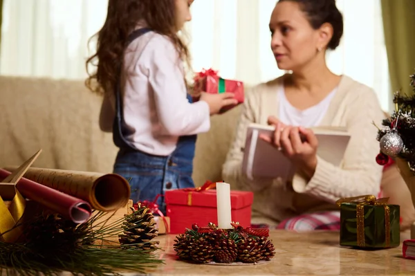 Bir muma, ambalaj malzemelerine ve güzel bir çocuğun bulanık arka planına odaklan, mavi kot tulumlu sevimli bebek annesine sevimli bir Noel hediyesi verir. Yeni Yıl