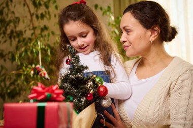 Güzel bir genç kadın, sevgi dolu bir anne küçük bir Noel ağacına oyuncak toplar ve takılar asan sevimli küçük kızına sarılıyor ve yeni yılı kutlamak için hazırlık yapıyor.