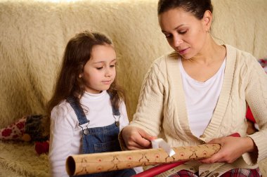 Mutlu bir anne ve kızı dekoratif bir kurdele seçip geyik desenli ambalaj kağıdının üzerine koyuyorlar. Yılbaşı hediyelerini ve hediye kutularını paketliyorlar. Kış etkinliklerinin beklentisinde