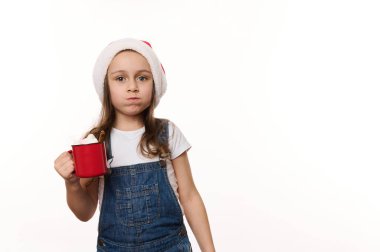 Yaramaz 5 yaşında Noel Baba şapkası ve kot pantolon giyen sıcak çikolata içen ve marshmallow yiyen, beyaz arka planda izole edilmiş reklam alanı olan küçük bir kız çocuğu. Noel atmosferi