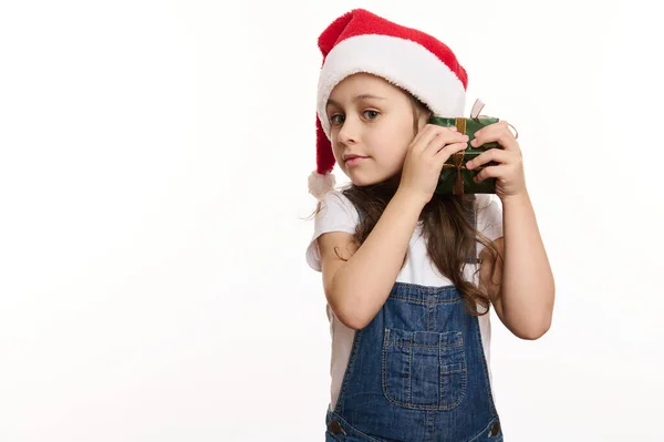 Isolated portrait on white background of a happy, adorable curious little child girl in Santa hat, shaking a Christmas present and guessing whats inside. Copy ad space. Time to open Xmas gift boxes