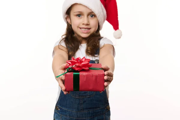 Portrait on white background of a lovely little girl in Santa hat, handing a happy gift box with beautiful tied red bow, cutely smiles looking at camera. Christmas Eve. New Year and winter holidays.