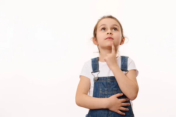 stock image Beautiful pensive little girl wearing blue denim overalls, holding her finger on her chin, thoughtfully looking up at a copy advertising space for promotional text, isolated over white background