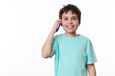 Coşkulu, koyu saçlı, turkuaz tişört giyen, neşeli, dişlek bir gülümseme takınan, cep telefonuyla konuşurken kameraya bakan, beyaz arka planda fotokopi çeken beyaz çocuk.